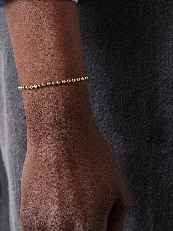 Gold beaded bracelet on a wrist against a dark textured background