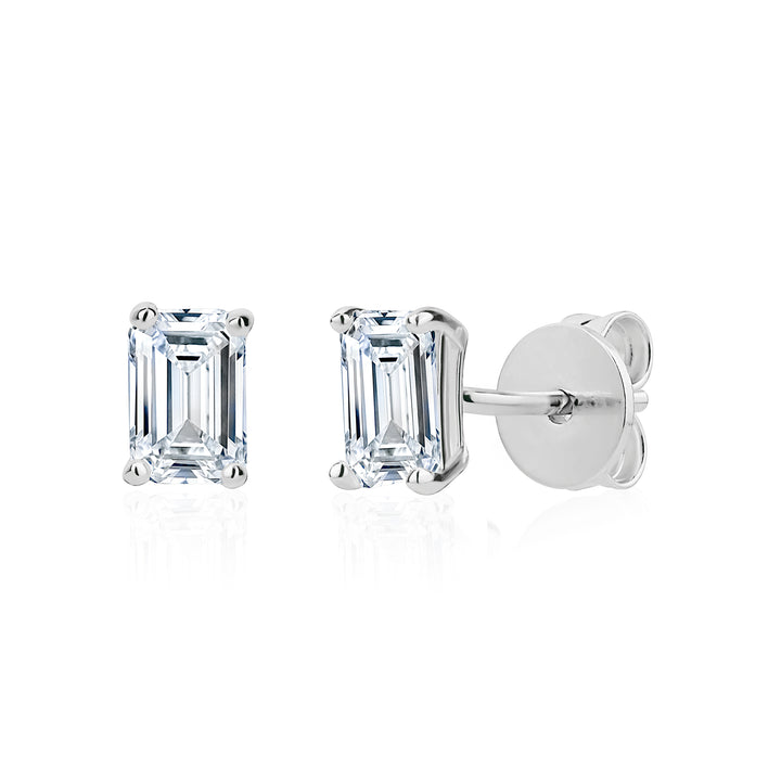 A pair of emerald-cut diamond stud earrings in a silver setting, with one earring facing forward and the other angled to show the post and butterfly back, on a white background with a soft reflection.
