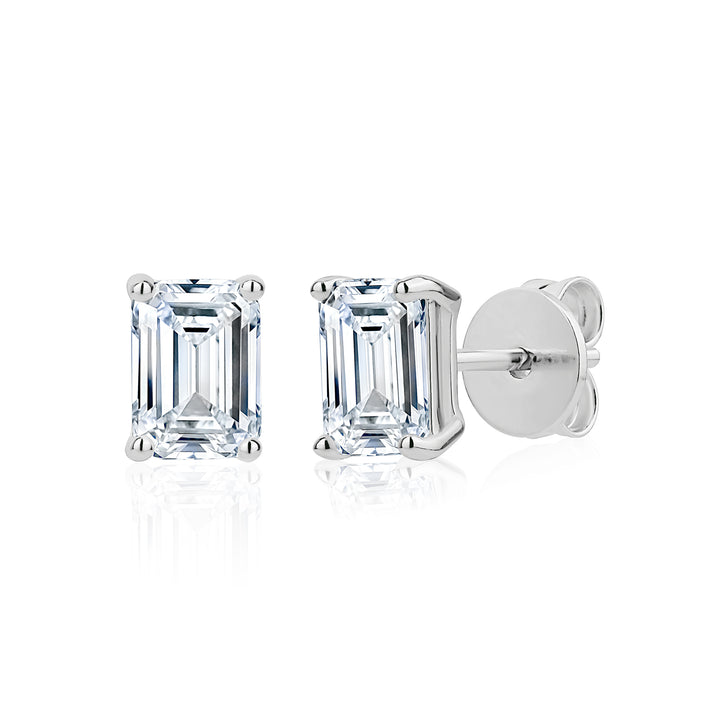 A pair of emerald-cut diamond stud earrings in silver settings, showing both the front view of the clear gemstone and the side profile with the post and butterfly back, on a white background with a soft reflection.