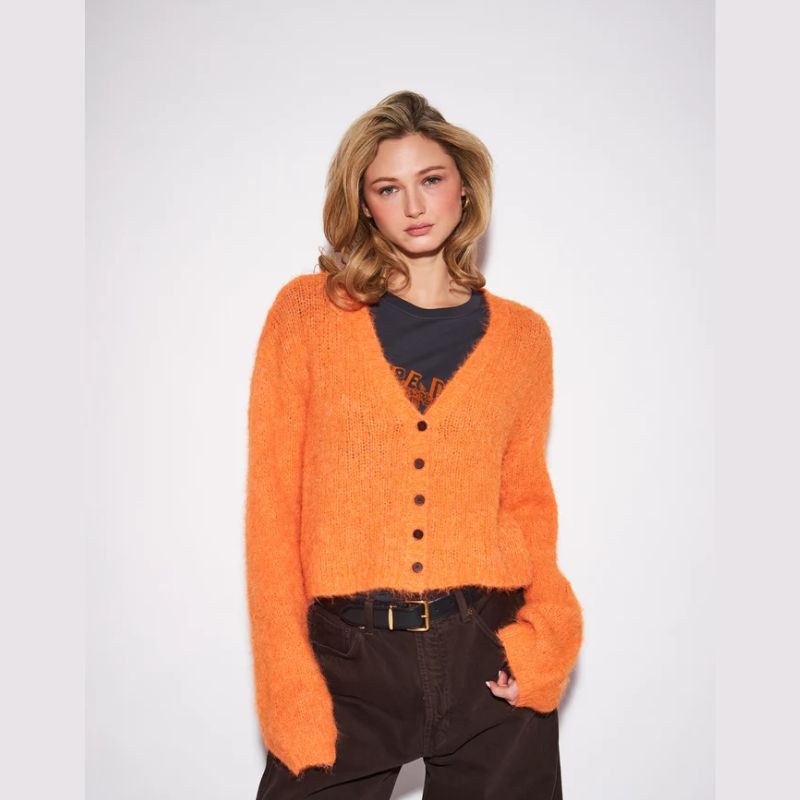 A woman modeling a fuzzy, textured orange cardigan sweater with dark buttons, worn over a black graphic t-shirt and dark brown pants, against a plain light gray background.