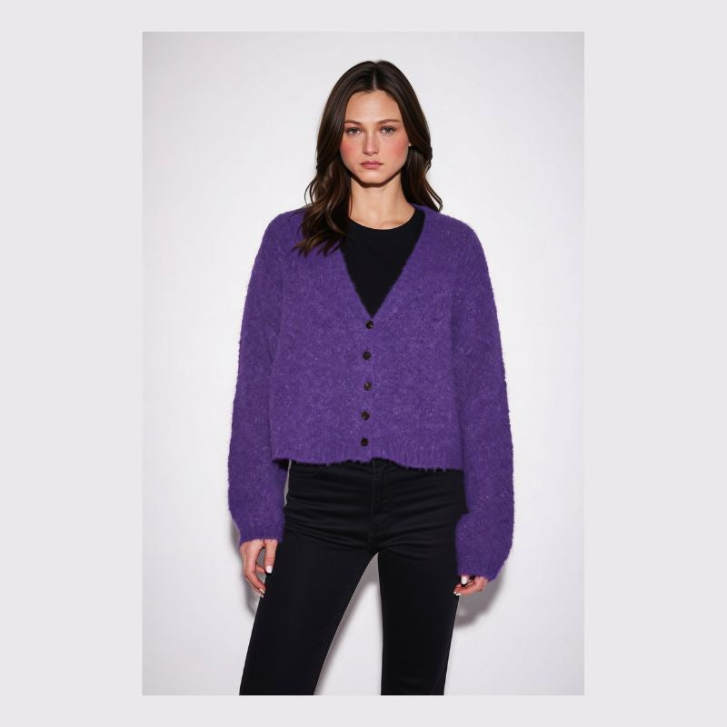 A woman modeling a fuzzy purple button-front cardigan over a black top and black pants, photographed against a plain light gray studio background.