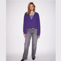 A model stands against a light gray studio background wearing a fuzzy purple button-front cardigan over a gray graphic T-shirt, gray jeans, and black pointed shoes.