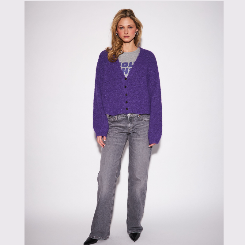 A model stands against a light gray studio background wearing a fuzzy purple button-front cardigan over a gray graphic T-shirt, gray jeans, and black pointed shoes.
