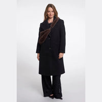 A woman modeling a long black coat with a dark brown faux-fur collar, paired with black lace-up pants, against a plain light gray studio background.
