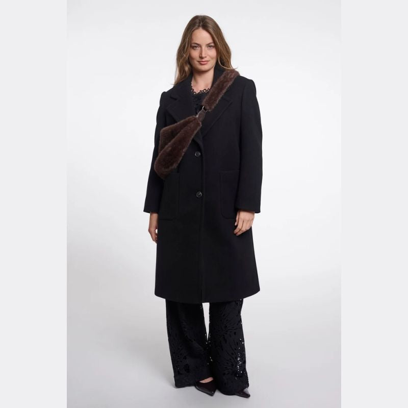 A woman modeling a long black coat with a dark brown faux-fur collar, paired with black lace-up pants, against a plain light gray studio background.