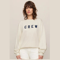 A woman wearing a cream-colored crewneck sweatshirt with the word 'CREW' printed in navy blue across the chest, paired with matching cream shorts, set against a solid light beige background.
