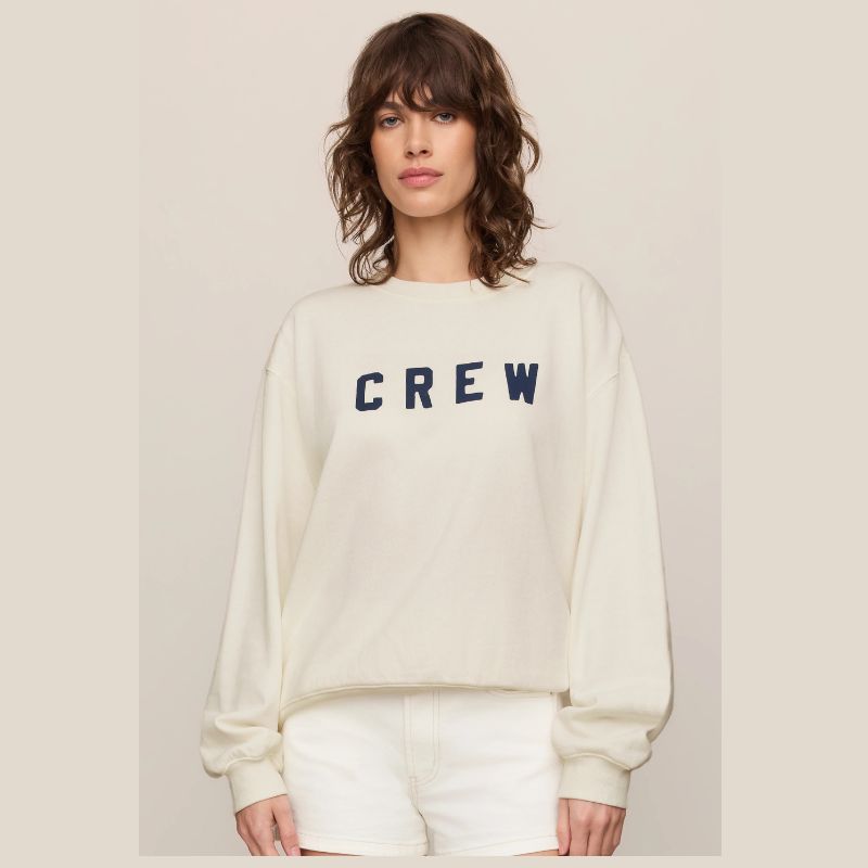 A woman wearing a cream-colored crewneck sweatshirt with the word 'CREW' printed in navy blue across the chest, paired with matching cream shorts, set against a solid light beige background.