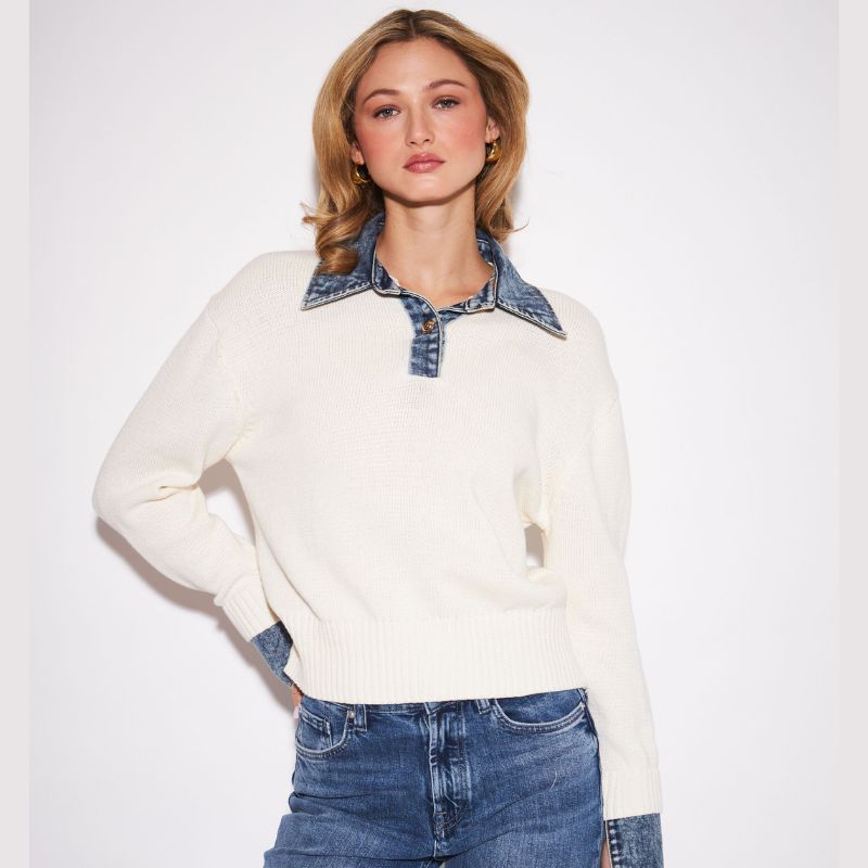 A woman modeling a cream knit sweater with a denim collar and matching denim cuffs, paired with blue jeans, against a plain white studio background.
