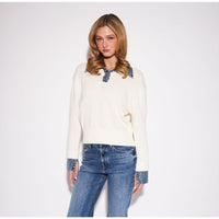 A woman modeling a cream ribbed knit sweater with blue denim collar and cuff details, paired with blue jeans, against a plain white studio background.