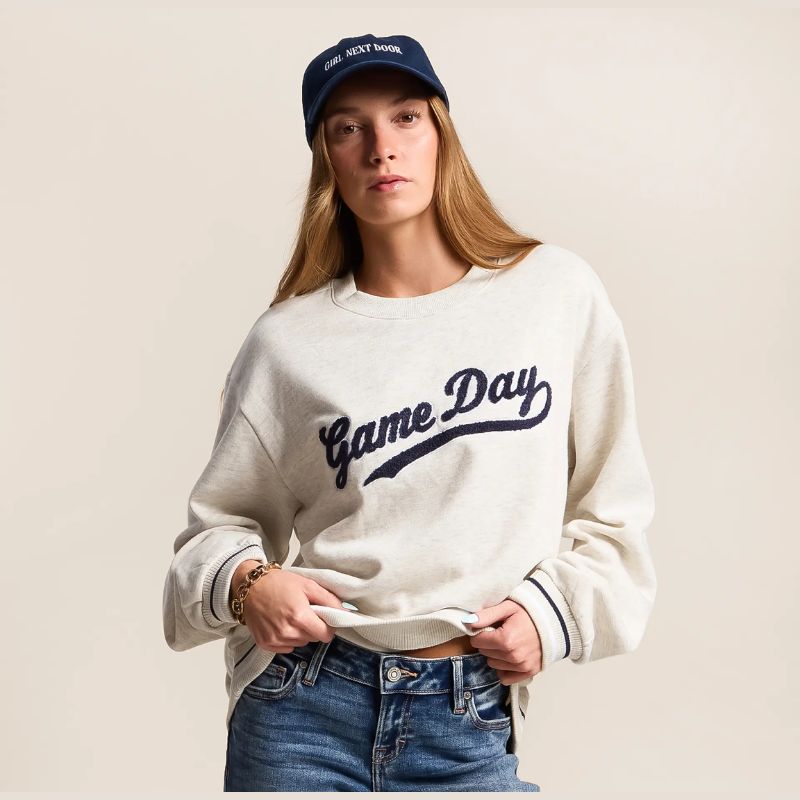 A woman wearing a cream-colored crewneck sweatshirt with 'Game Day' embroidered in navy blue script across the chest, paired with blue jeans and a navy blue baseball cap.