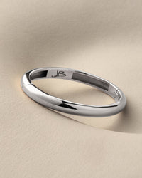 A polished silver band ring with a smooth, rounded profile, resting on a textured beige surface. The inside of the band features a custom engraving of the initials 'JB'.