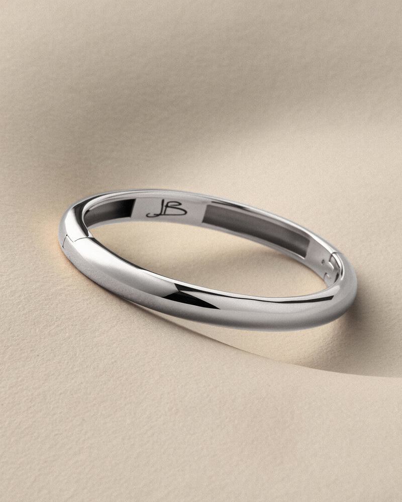 A polished silver band ring with a smooth, rounded profile, resting on a textured beige surface. The inside of the band features a custom engraving of the initials 'JB'.