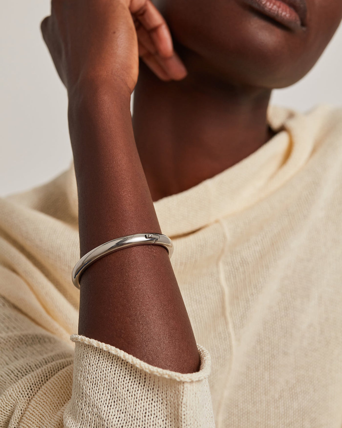 A close-up of a model wearing a polished silver bangle bracelet on her wrist, paired with a cream-colored knit top against a neutral light background.