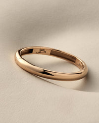 A polished rose gold band ring with a smooth, rounded profile, featuring an engraved monogram 'JB' inside the band, resting on a textured beige surface.