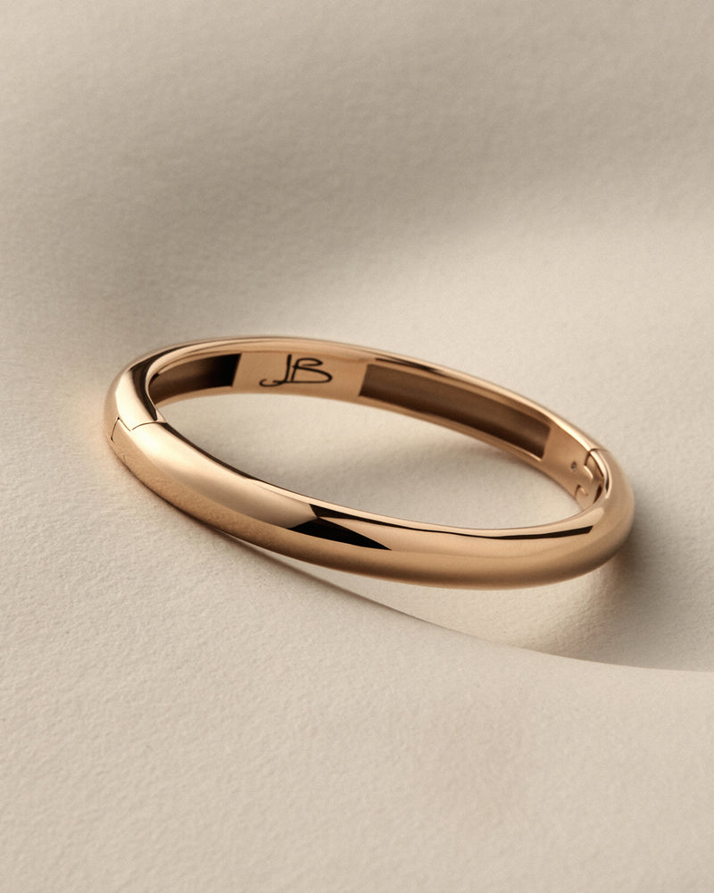 A polished rose gold band ring with a smooth, rounded profile, featuring an engraved monogram 'JB' inside the band, resting on a textured beige surface.