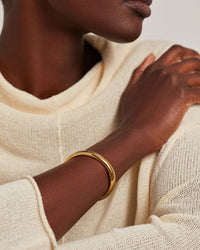 A close-up of a person wearing a polished gold bangle bracelet on their wrist, dressed in a cream-colored knit sweater against a neutral light background.