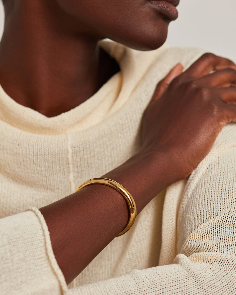 A close-up of a person wearing a polished gold bangle bracelet on their wrist, dressed in a cream-colored knit sweater against a neutral light background.