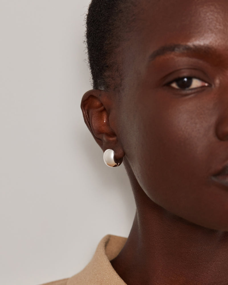 A close-up of a model's ear wearing a small, round, white pearl-like stud earring with a subtle gold-tone rim, set against a neutral light gray background.