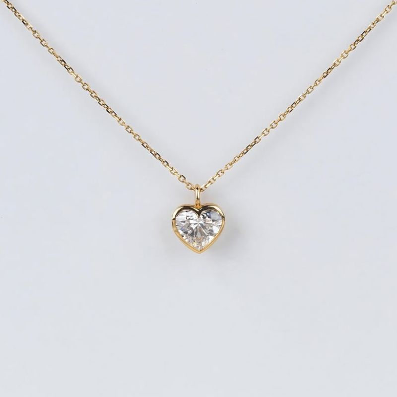 A delicate gold chain necklace with a small heart-shaped pendant featuring a clear heart gemstone in a gold bezel setting, displayed against a light gray background.