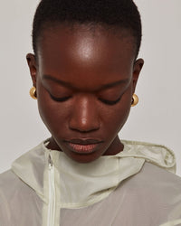Close-up of a model wearing gold hoop earrings and a light beige hooded windbreaker jacket against a neutral studio background.