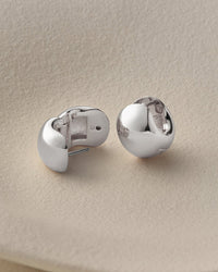 A pair of polished silver stud earrings with a rounded, organic shape, displayed on a textured beige surface.