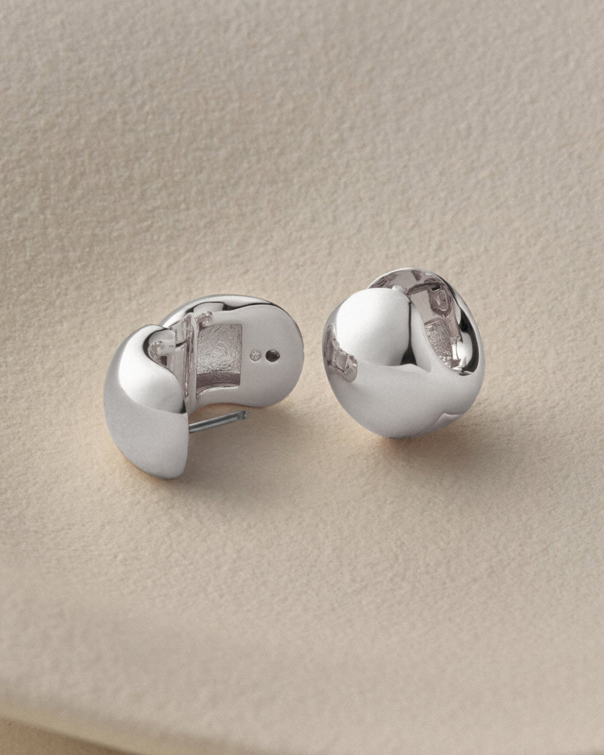 A pair of polished silver stud earrings with a rounded, organic shape, displayed on a textured beige surface.