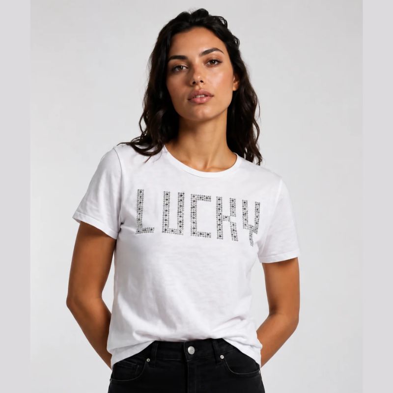 A woman wearing a white short-sleeve t-shirt with LUCKY spelled out in silver  studs across the chest, paired with black jeans, against a light gray studio background.