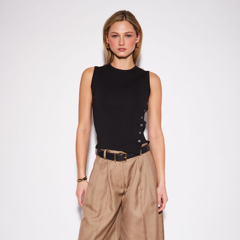 A woman modeling a sleeveless black knit top with side buttons, paired with high-waisted tan trousers and a black belt, against a plain white studio background.