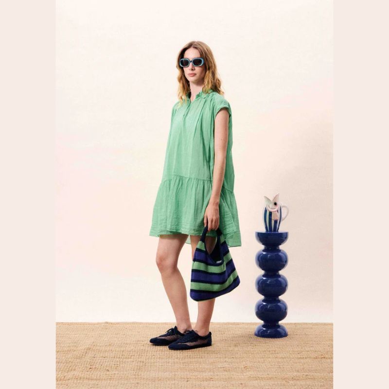 Woman in a short sleeve green cotton  dress standing next to a blue and green striped bag on a beige floor.