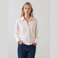 A woman modeling a light pink long-sleeve button-up shirt with dark blue jeans, standing against a plain light gray background.