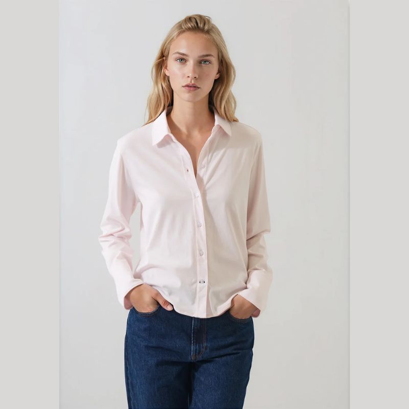 A woman modeling a light pink long-sleeve button-up shirt with dark blue jeans, standing against a plain light gray background.