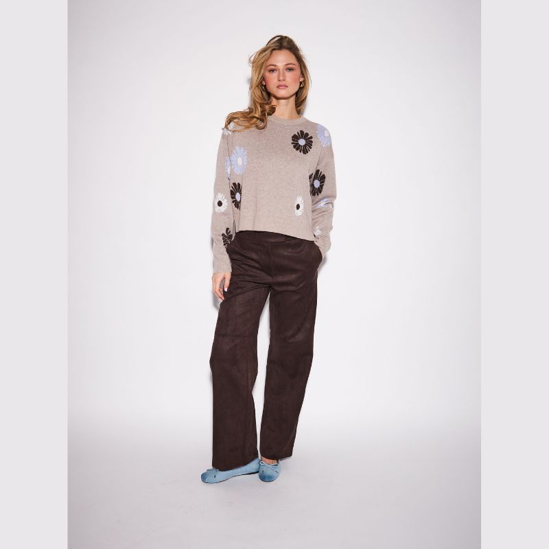 A woman modeling a beige knit sweater with dark brown and light blue floral patterns, paired with dark brown wide-leg trousers and light blue slip-on shoes, against a light gray studio background.