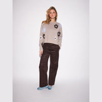 A woman modeling a beige knit sweater with dark brown and light blue floral patterns, paired with dark brown wide-leg trousers and light blue slip-on shoes, against a light gray studio background.