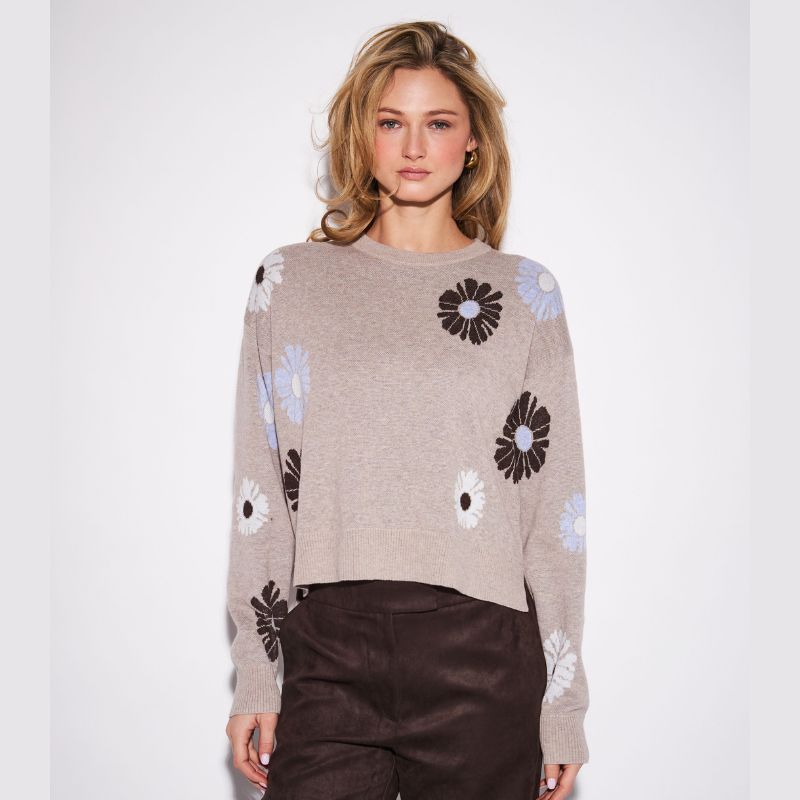 A woman modeling a cropped knit sweater in taupe with large black, white, and light blue flower motifs, paired with dark brown pants, against a plain light gray studio background.
