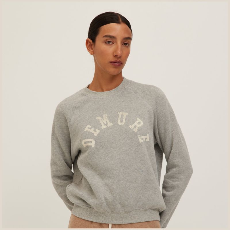 A model wearing a light gray heather crewneck sweatshirt with the word 'DEMURE' arched in cream collegiate lettering across the chest, set against a plain light gray background.