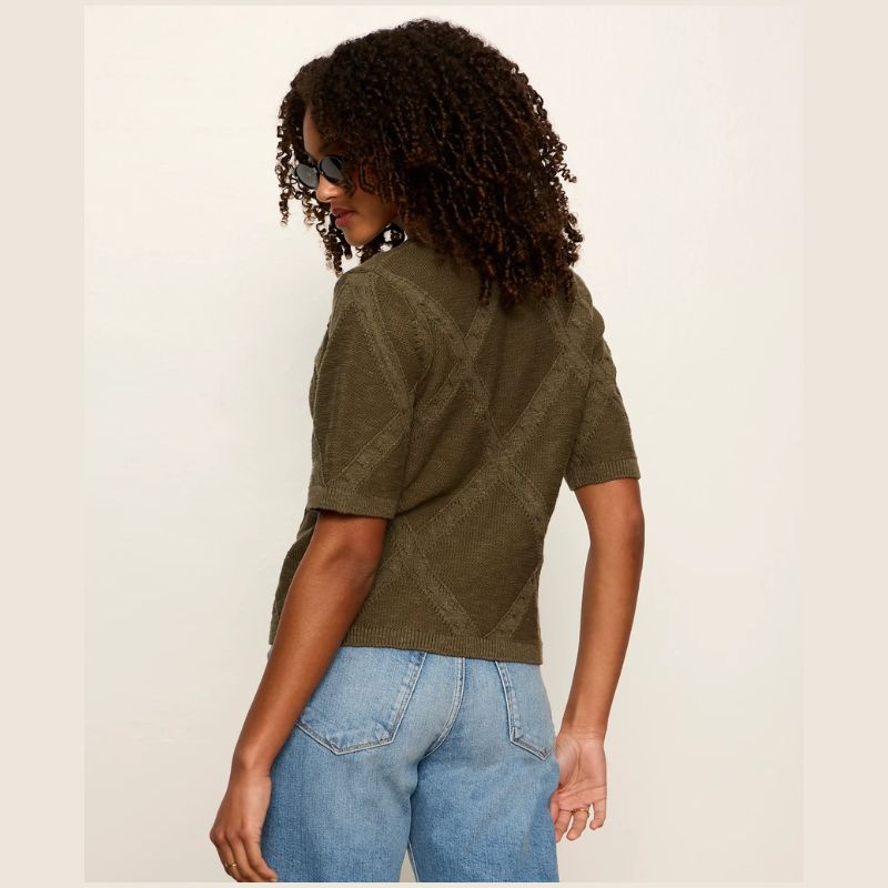 A woman with curly hair wearing a short-sleeve olive green knit top and light blue jeans, shown from a side-back angle against a plain off-white background.