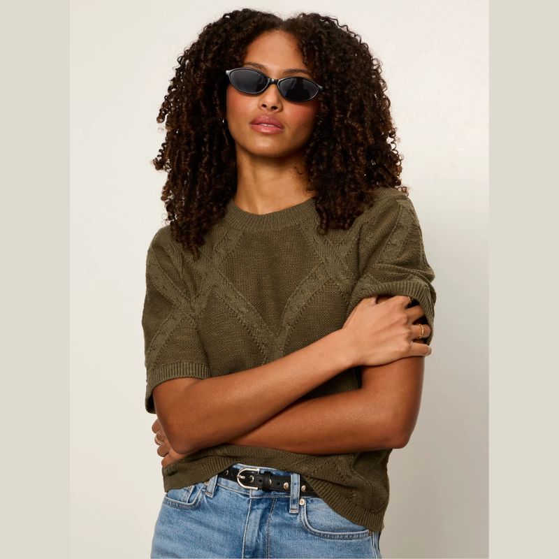 A woman with curly dark hair wearing a short-sleeve cotton Kalamata olive green knit sweater, blue jeans, and black sunglasses, posed with her arms crossed against a plai