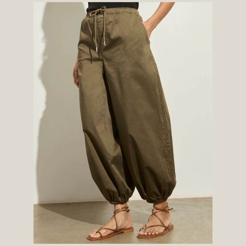 Woman wearing a black tank top and Kalamata olive green poplin  sateen pants with elastic cuff at hem against a plain background