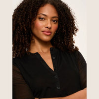 A woman with curly dark hair wearing a black short-sleeve henley top with sheer mesh sleeves, shown from the chest up against a plain light background.