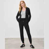 A woman modeling a black zip-up hoodie and matching black jogger sweatpants, styled with a gray tank top and black sneakers, in a minimalist studio setting.