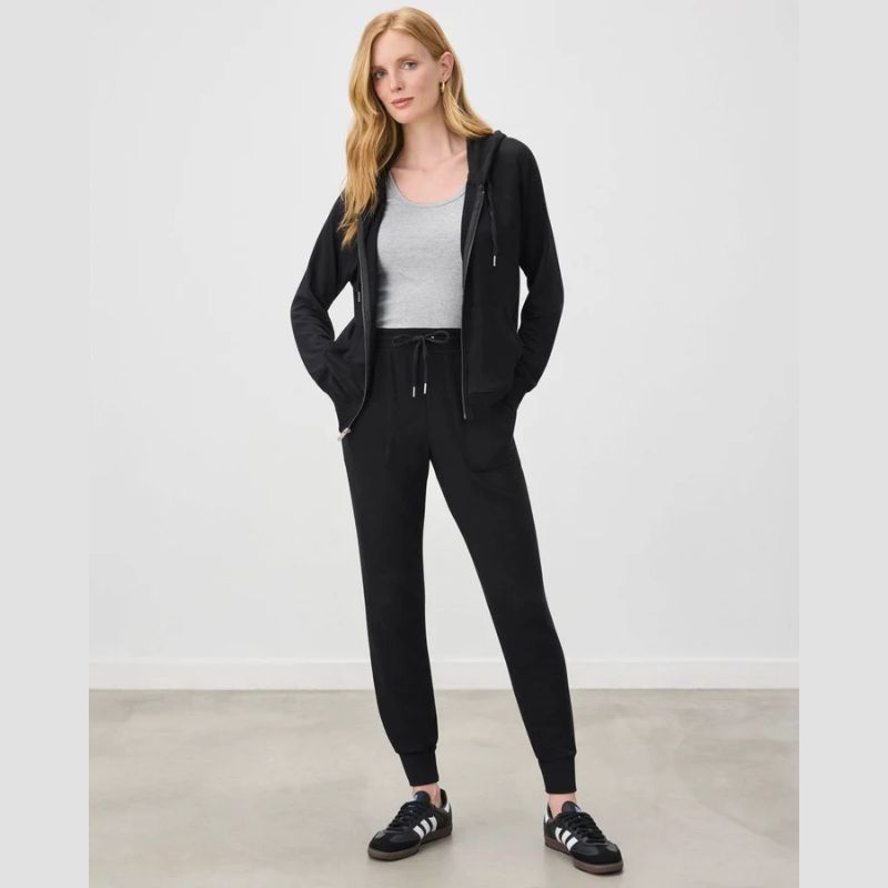 A woman modeling a black zip-up hoodie and matching black jogger sweatpants, styled with a gray tank top and black sneakers, in a minimalist studio setting.