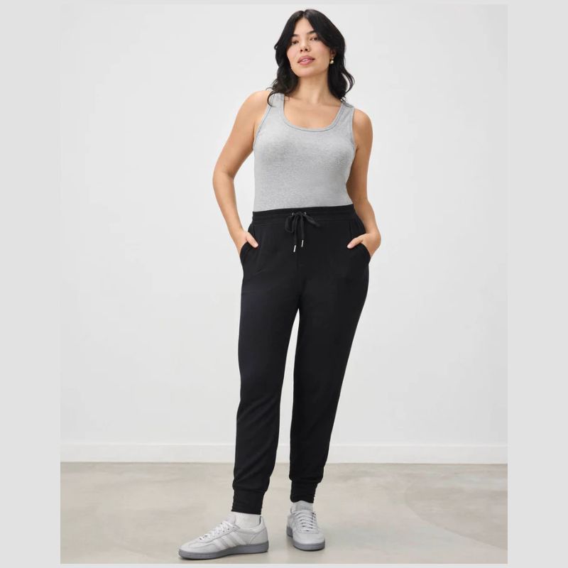 A woman modeling a light gray ribbed tank top and black jogger sweatpants with white sneakers, standing against a plain light gray studio wall.