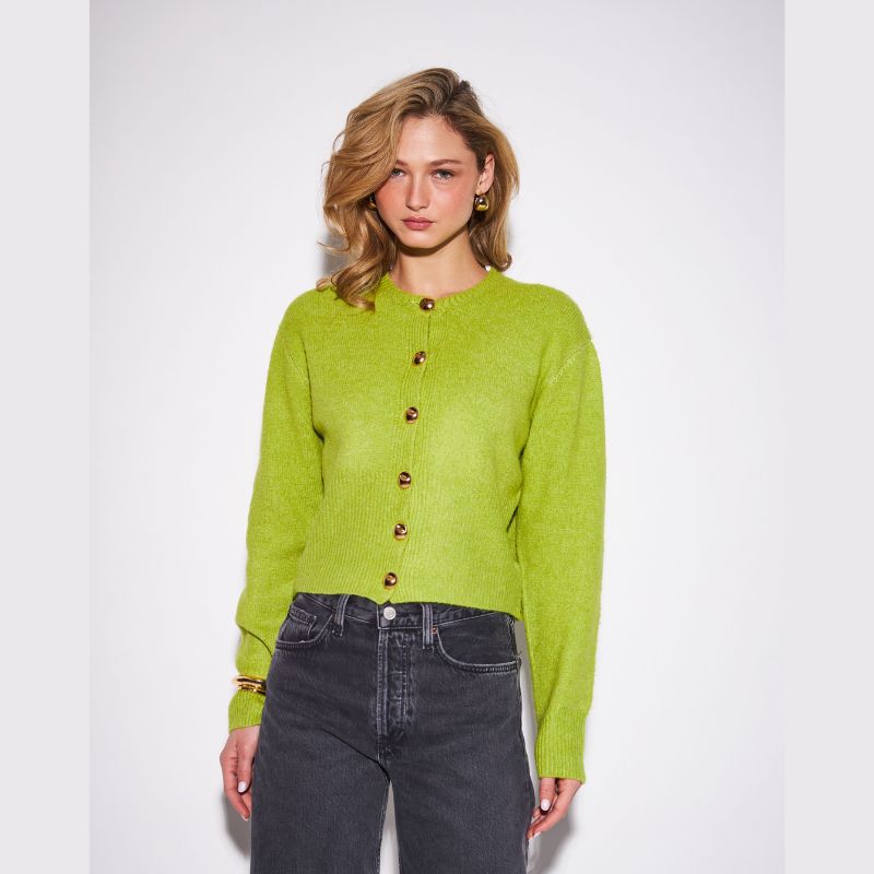 A woman modeling a lime green cropped button-up cardigan sweater with dark gray jeans, standing against a light gray studio background.