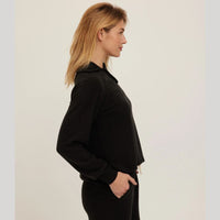 A woman in profile wearing a black long-sleeve cropped zip-up hoodie and black pants, standing against a plain light gray studio background.