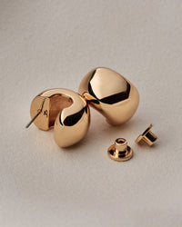 A pair of polished gold-toned geometric stud earrings with a matching flat back, displayed on a textured light beige surface.