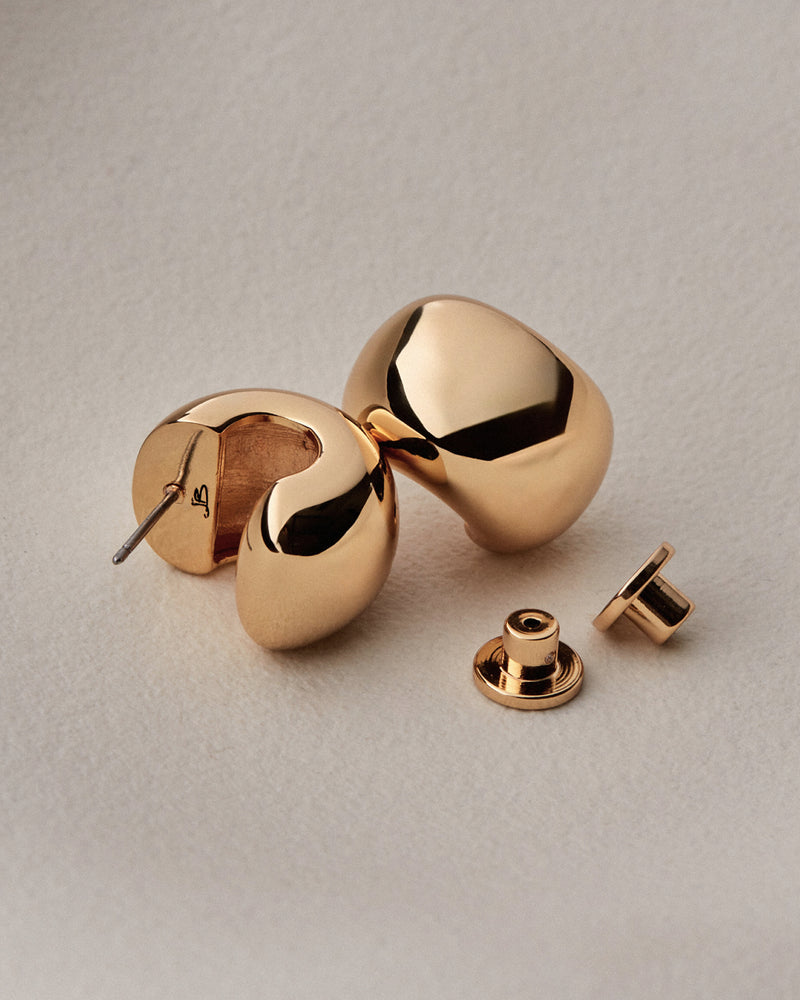A pair of polished gold-toned geometric stud earrings with a matching flat back, displayed on a textured light beige surface.