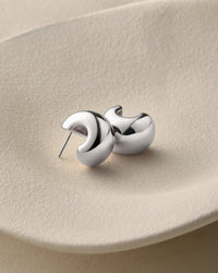 A pair of polished silver crescent-shaped stud earrings resting on a textured beige ceramic plate.