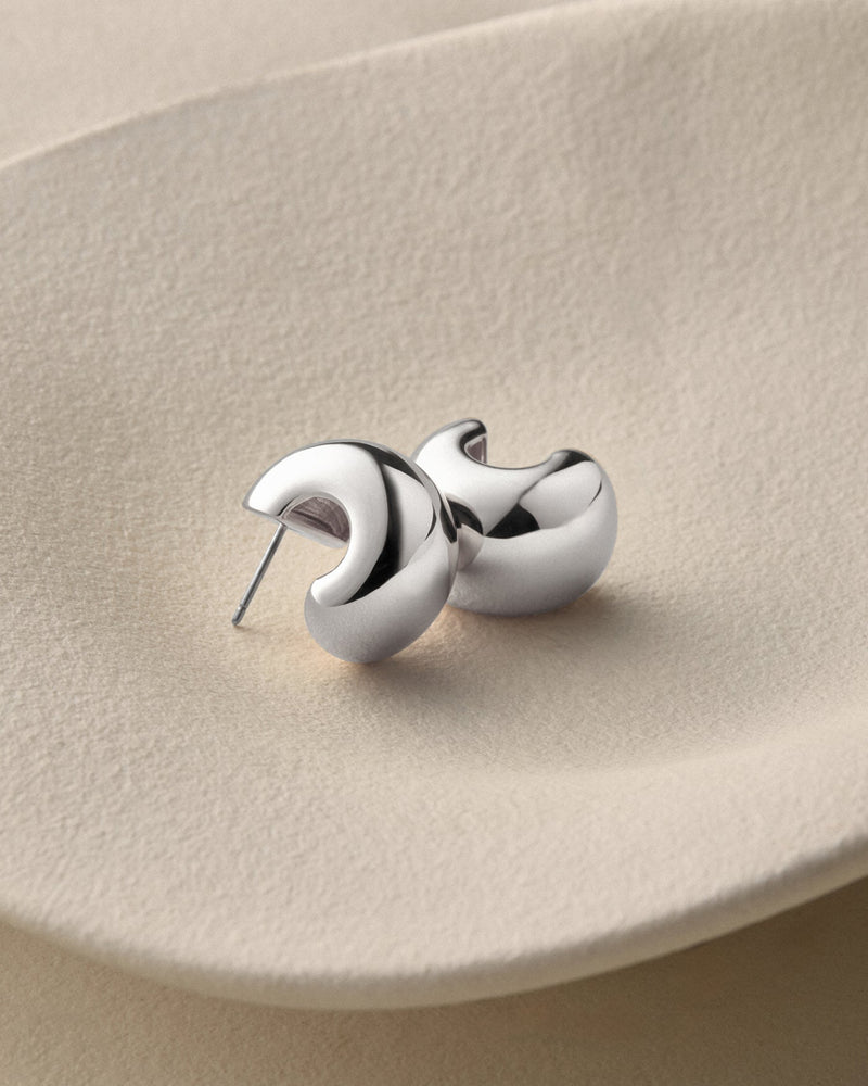A pair of polished silver crescent-shaped stud earrings resting on a textured beige ceramic plate.