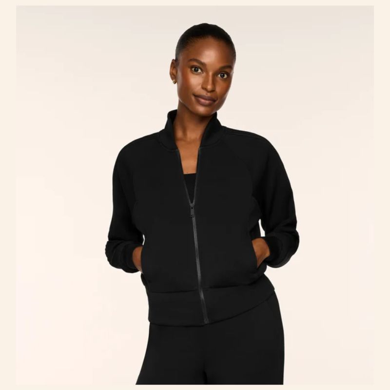 A woman modeling a black zip-up athletic jacket with a stand collar and side pockets, paired with black pants, against a solid light beige background.