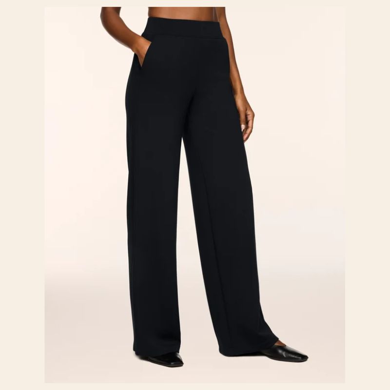 A model wearing black wide-leg trousers with side pockets, paired with black pointed flats, shown from the waist down against a light beige background.